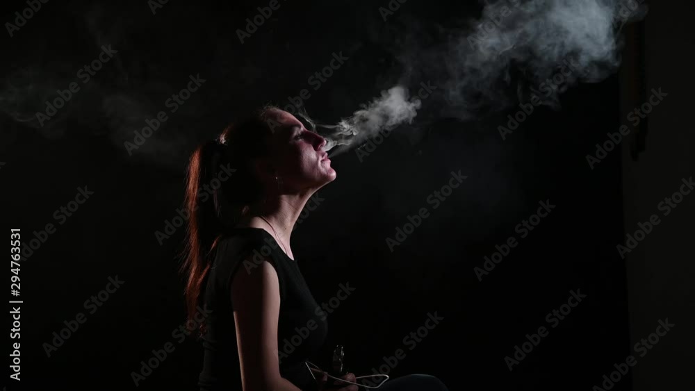 The brunette smokes an electronic cigarette on a black background, releases thick smoke from her mouth. Portrait of a woman smoking a vape, hovering. The brunette smokes an electronic cigarette on a black background, releases thick smoke from her mouth. Portrait of a woman smoking a vape, hovering.