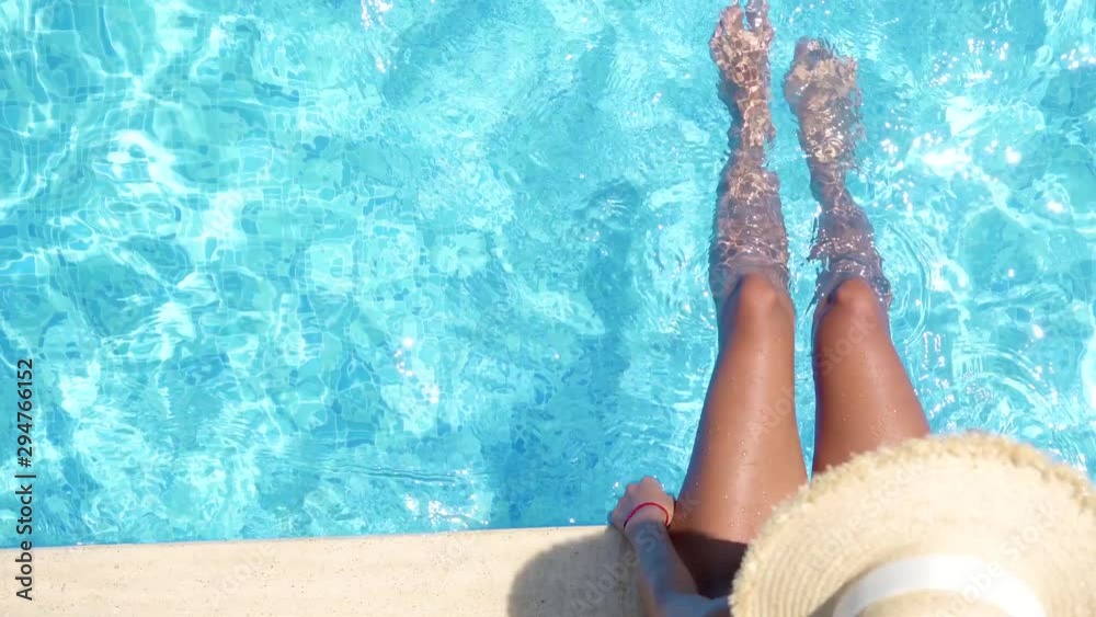 Girl in bikini relaxing in swimming pool in sunny day. Beautiful sexy girl in summer pool splashing water with feet. Top view of female beautiful legs in water. 4K