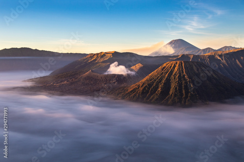 Mount Bromo volcano at sunrise; Island of East Java, Indonesia. Clouds cover the valley floor; Luhur Poten Temple at the foot of the cone. Gas from another volcano rising against the