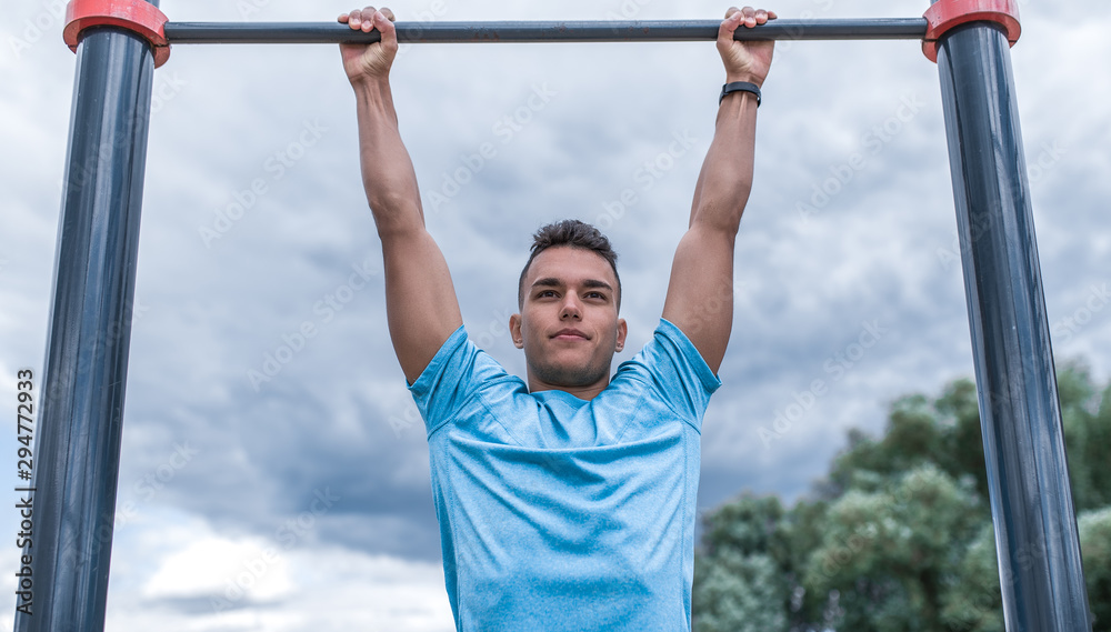 Naklejka premium sportowiec podciąga się na pasku. Latem wiosną w mieście. Aktywny tryb życia, trening, fitness na świeżym powietrzu. Motywacja do sportu. Niebieska koszulka, chmury w tle.