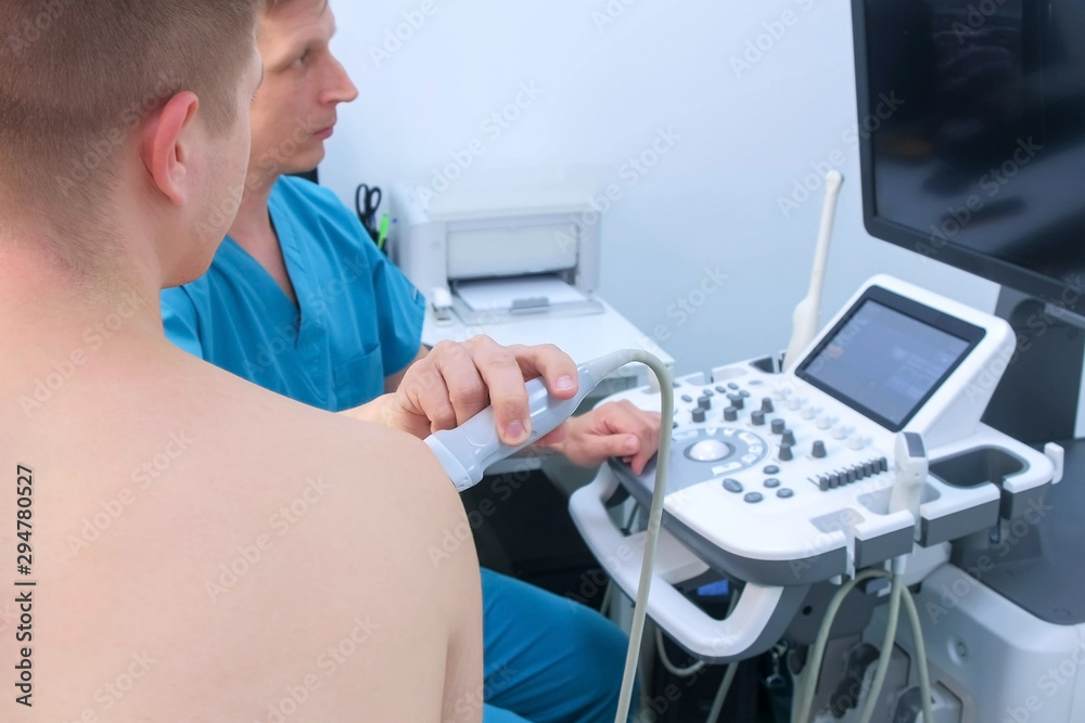 Doctor makes ultrasound of shoulder joint for man using ultrasound scanner in clinic. He runs ultrasound sensor over man's arm, working on scanner panel and looking at screen. Doctor makes ultrasound of shoulder joint for man using ultrasound scanner in clinic. He runs ultrasound sensor over man's arm, working on scanner panel and looking at screen.