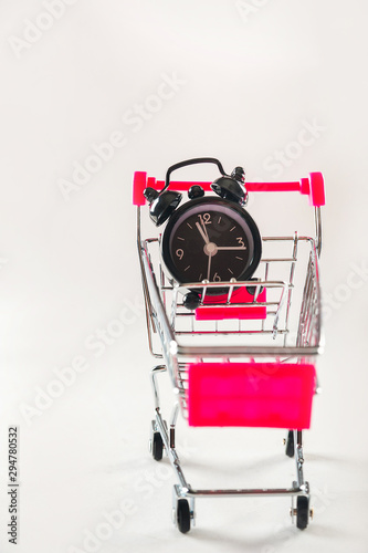Small shopping cart with alarm clock isolated on white background. Alarm clock in the miniature shopping trolley. The concept of time and shopping.