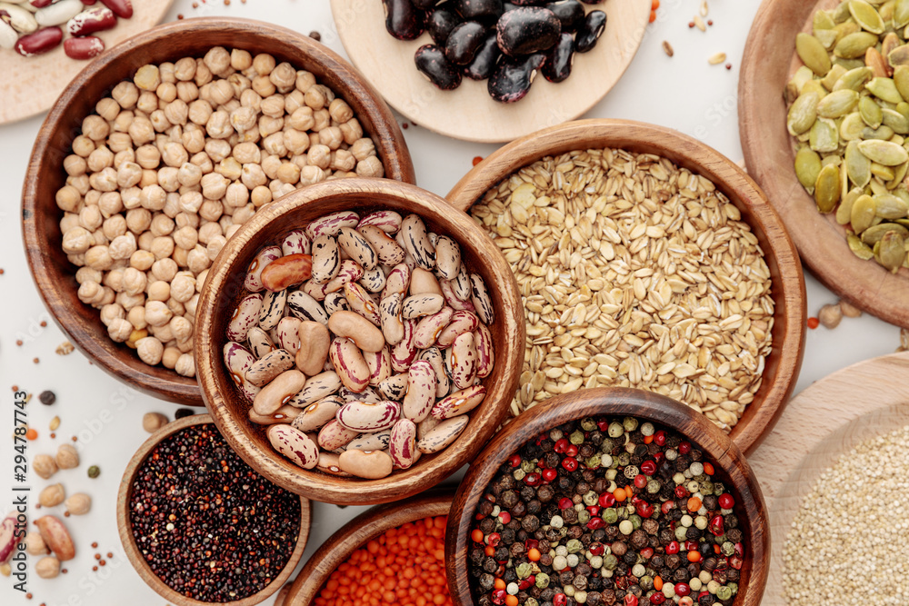 top view of wooden bowls with diverse beans, peppercorns, pumpkin seeds, oatmeal, quinoa and chickpea on white marble surface with scattered grains