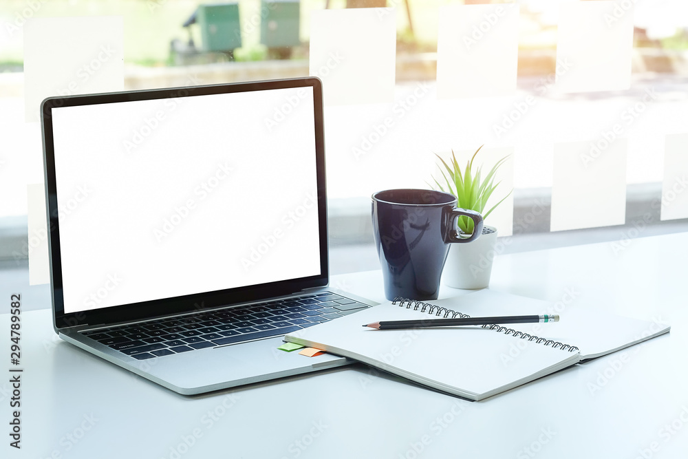 Fototapeta premium Computer laptop, coffee cup, potted plant and notebook on white office desk near glass window.