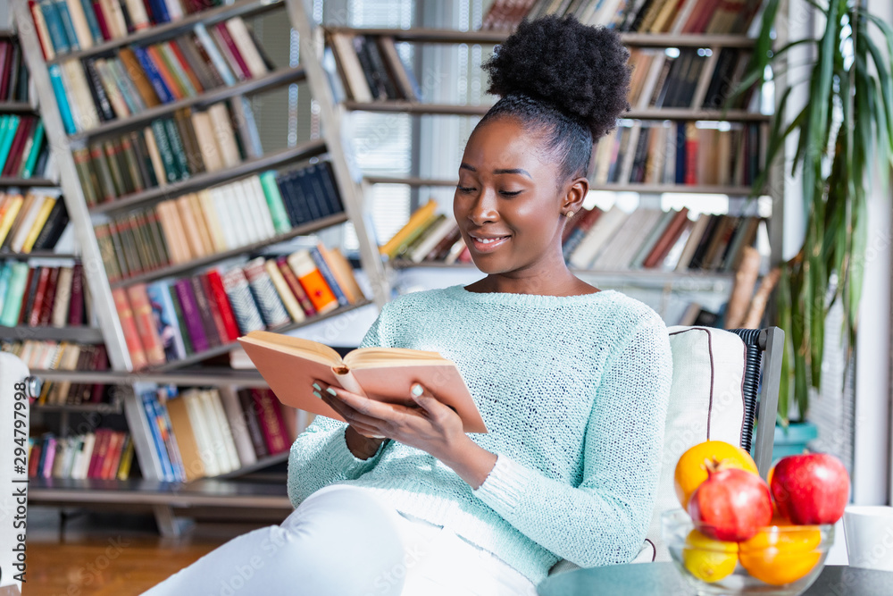 Φωτογραφία Young beautiful African American girl reading a book on the couch with the library bookshelves in the back