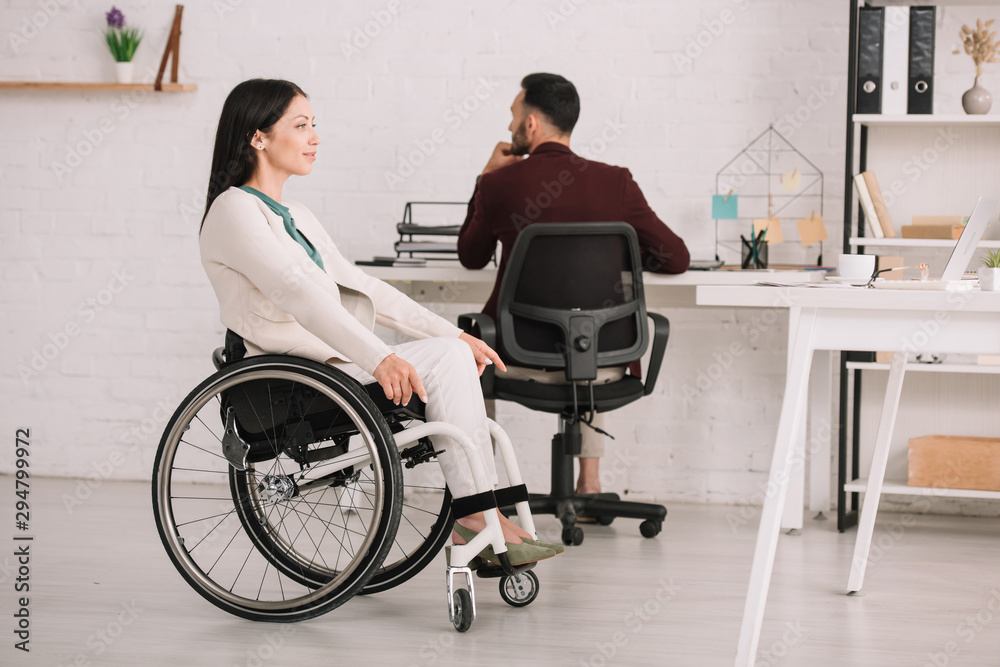 smiling disabled businesswoman looking away while sitting in wheelchair near colleague smiling disabled businesswoman looking away while sitting in wheelchair near colleague