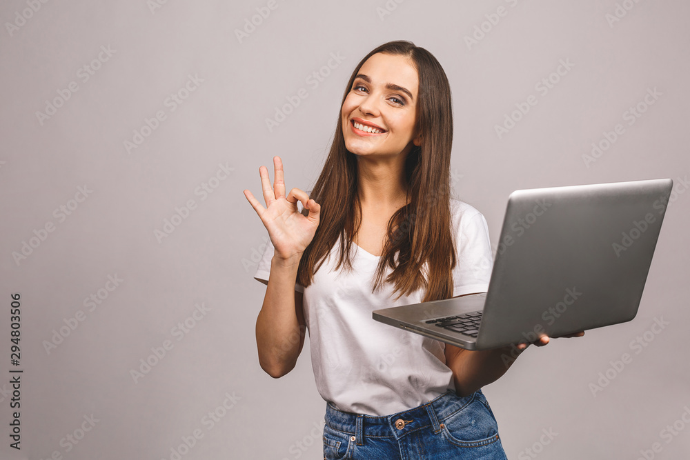 Portrait of a smiling young girl holding laptop computer and showing ok gesture isolated over grey background. - obrazy, fototapety, plakaty 