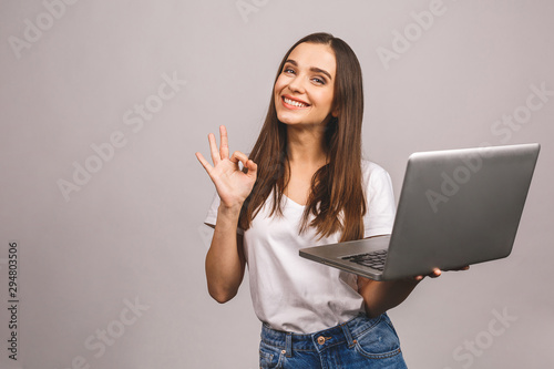 Portrait of a smiling young girl holding laptop computer and showing ok gesture isolated over grey background.