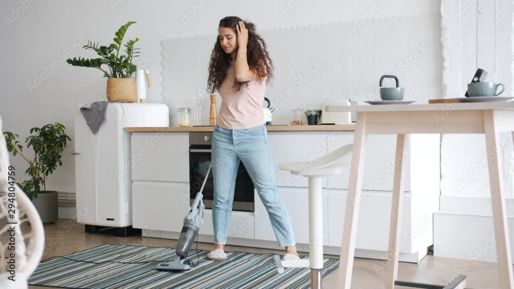 Happy young woman is vacuuming carpet in kitchen dancing having fun during clean-up in apartment. Housekeeping, lifestyle and creative people concept. Happy young woman is vacuuming carpet in kitchen dancing having fun during clean-up in apartment. Housekeeping, lifestyle and creative people concept.