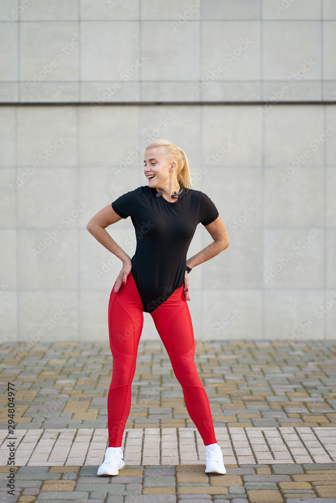 Sport and health. Attractive happy slim girl wearing black shirt, stylish red  leggings and white sneakers posing on grey wall background. Stock Photo