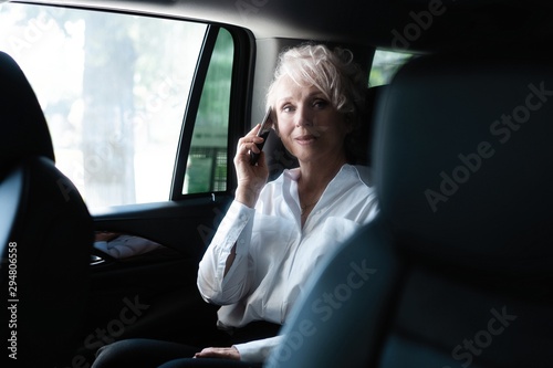 Cheerful mature woman in business suit sitting on backseat of her car and talking on mobile phone. © opolja