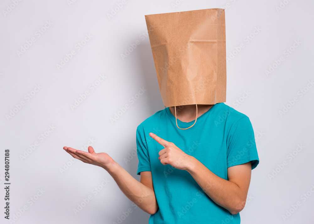 Teen boy with paper bag over head pointing finger away at copyspace, on