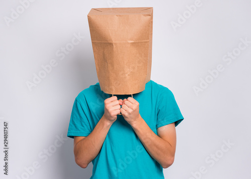 Portrait of teen boy with paper bag over head. Teenager cover head with bag holding hand near face posing in studio. Child pulling paper bag over head.