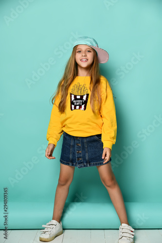 Canvas Print Funny teenage girl in a baseball cap and a yellow hoodie and a denim miniskirt posing in full growth on a blue background
