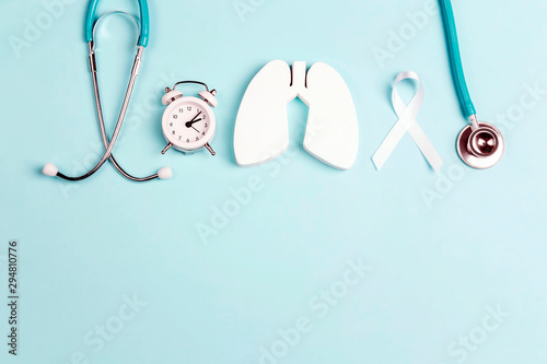 Lung cancer awareness background with white ribbon, alarm clock and stethoscope on blue.