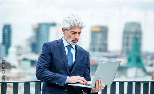 A mature businessman with laptop standing on a terrace, working.