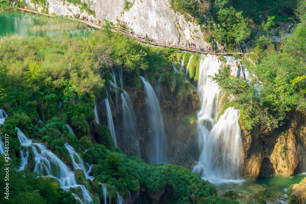 Fototapeta premium Tourists walking along the wooden boardwalk above the Large Waterfall at the Plitvice Lakes National Park in Croatia