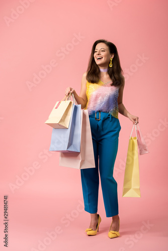 full length view of happy stylish disco girl holding shopping bags on pink