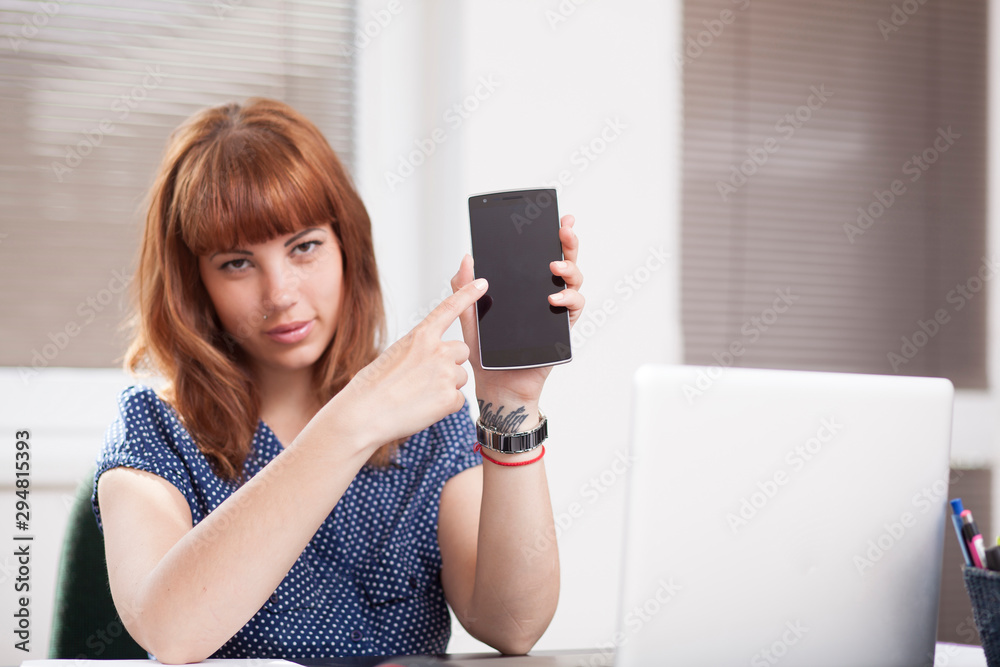 Naklejka premium Nice girl with tattoo and piercing showing a phone display (blank screen) and point at it with index finger, promoting app or website