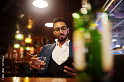 Handsome well-dressed arabian man with glass of whiskey and cigar hold mobile phone, posed at pub.