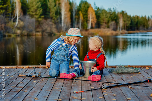 Two little sisters or friends sit with fishing rods on a wooden pier. They caught a fish and put it in a bucket. They are happy with their catch and discuss it.