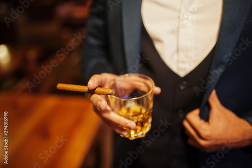 Close up hand of handsome well-dressed arabian man with glass of whiskey and cigar posed at pub.