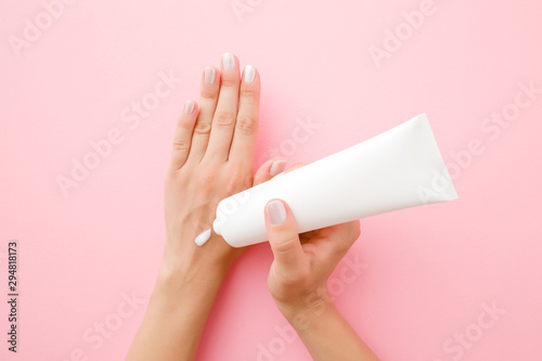 Young, perfect woman hands using moisturizing cream. Pastel pink background. Holding white tube. Care about clean, soft and smooth body skin. Point of view shot.