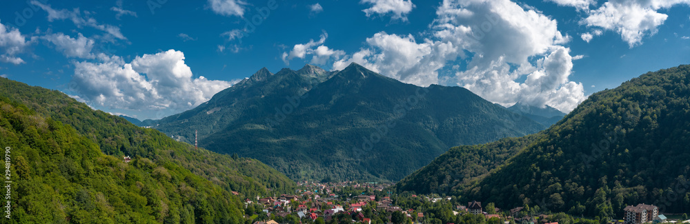 Panorama of Krasnaya Polyana mountain ski village, Aibga ridge in background; scenic picture-postcard view of rural terrain, alpine houses, beautiful light summer cloudy day, aerial drone view, Russia