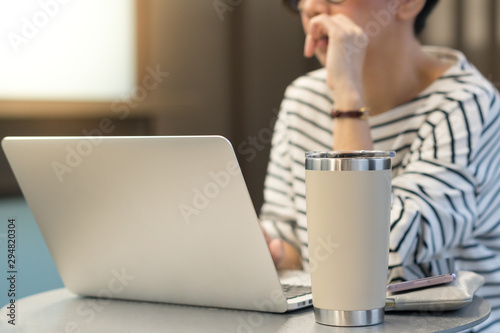 Smart looking woman use laptop computer to work from home due to Covid-19 pandemic, city lockdown and social distancing with her drink in a reusable stainless steel tumbler mug aside. photo
