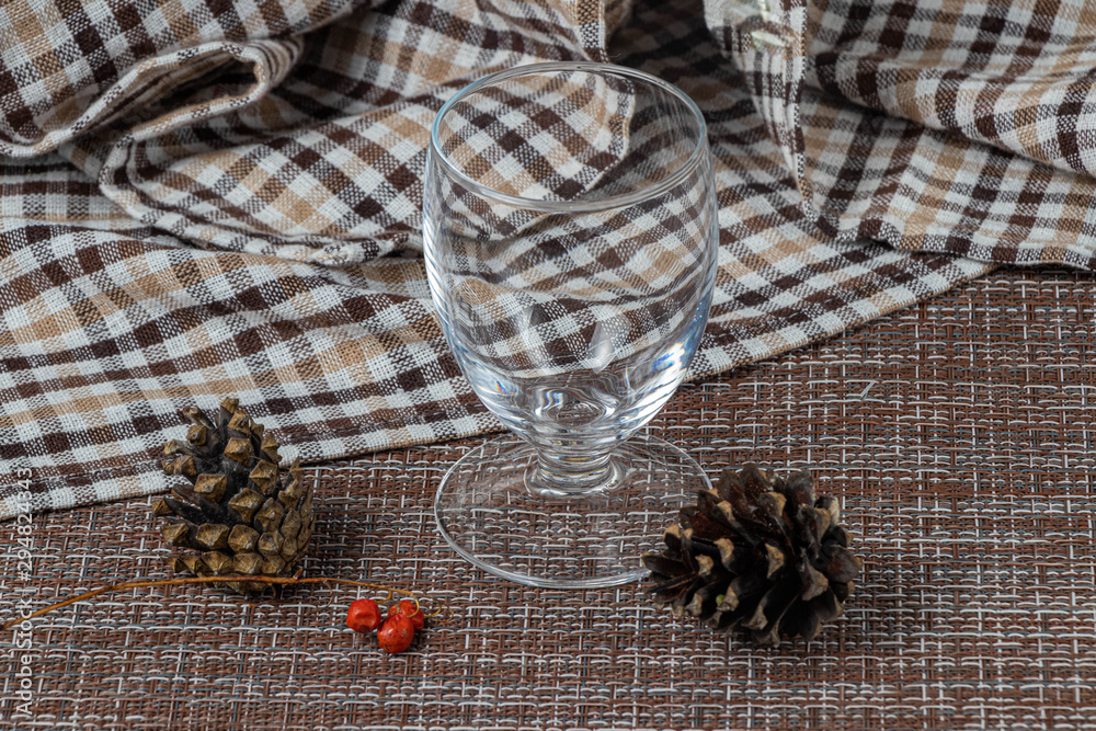 Empty transparent glass on a checkered white and brown kitchen towel. Spruce cones and red berries on a foreground. After the Christmas party. Empty transparent glass on a checkered white and brown kitchen towel. Spruce cones and red berries on a foreground. After the Christmas party.
