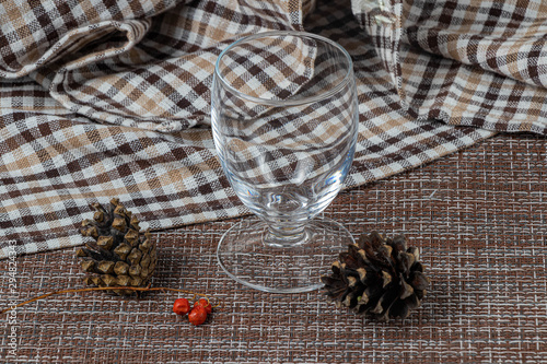 Fototapeta Naklejka Na Ścianę i Meble -  Empty transparent glass on a checkered white and brown kitchen towel. Spruce cones and red berries on a foreground. After the Christmas party.