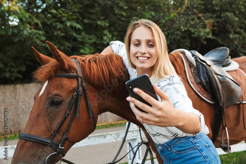 Beautiful smiling young blonde woman petting a horse at the stable
