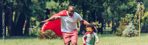 panoramic shot of african american father and son running in costumes of superheroes in park