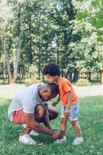 young african american man tying shoelaces of cute son in park