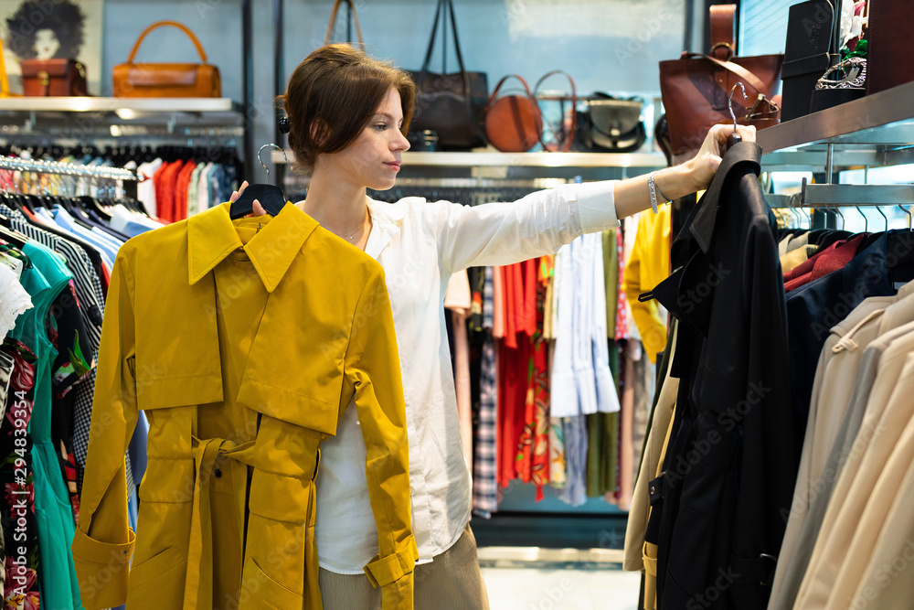 A young woman chooses between a black and mustard light coat in a clothing store.