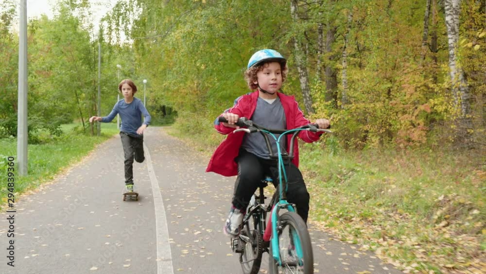 Teenage Boy Riding on Skateboard Brother on Cycle. Two Brothers Race at Autumn Park Pathway. Caucasian Kid with Helmet on Bicycle, Funny Friend Dashing on Board. Outdoor Sport Activity Teenage Boy Riding on Skateboard Brother on Cycle. Two Brothers Race at Autumn Park Pathway. Caucasian Kid with Helmet on Bicycle, Funny Friend Dashing on Board. Outdoor Sport Activity