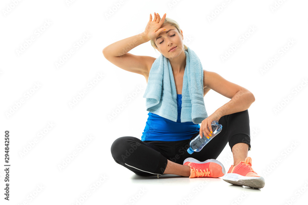 A young sports girl with blonde hair in a black sports axe, black leggings and bright sneakers with a towel around her neck and a bottle of water tired after training.