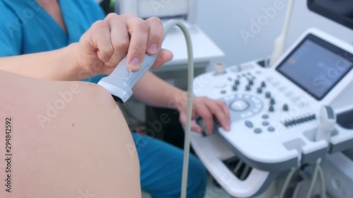 Doctor makes ultrasound of shoulder joint to man using ultrasound scanner in clinic, closeup. He runs ultrasound sensor over man's arm, working on scanner panel and looking at screen.