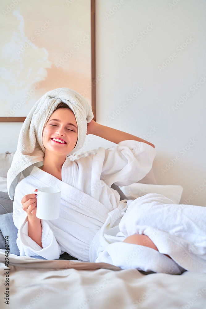 Photo of happy woman in white bathrobe with mug of tea in her hands lying on bed.