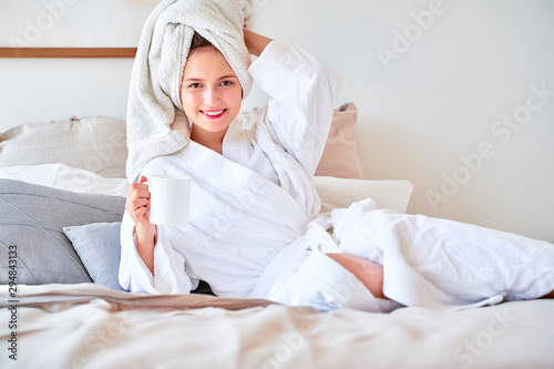 Image of young woman in white bathrobe with mug of tea in her hands lying on bed .