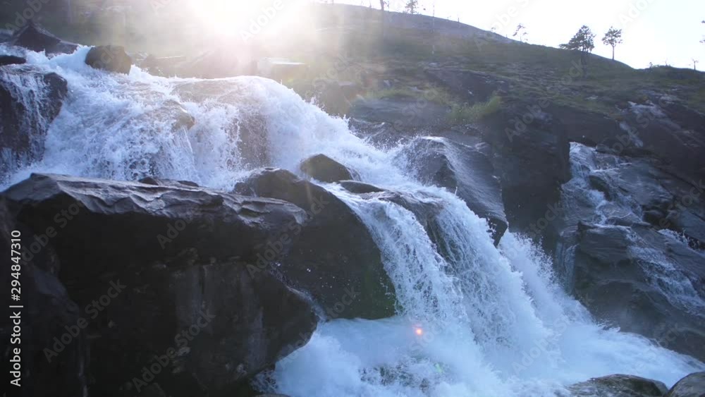 Static slow motion shot of a powerful and fast rapid going down at a steep angle, hitting big black rocks. Small waterfall as a part of a river rushing down. Sunshine peaks through behind the mountain Static slow motion shot of a powerful and fast rapid going down at a steep angle, hitting big black rocks. Small waterfall as a part of a river rushing down. Sunshine peaks through behind the mountain