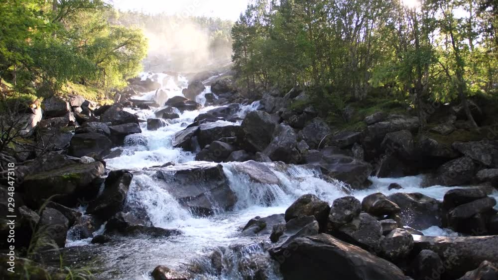 Static shot of a small peaceful river or stream, running down between rocks and lush green trees at sunset. Sunshine hitting the mist from the waterfall in the background.