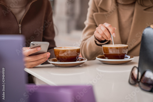 Cropped shot of young women using smartphone and drinking coffee after shopping at cafe outdoors