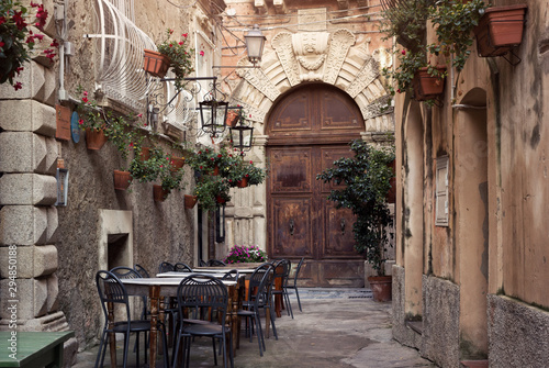 Fototapeta Naklejka Na Ścianę i Meble -  Tropea old city street with cozy restaurant terrace and floral decoration. Medieval wooden door of historical building and stone pavement.