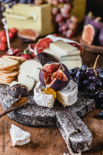 assortment of cheese on wooden board with fruits, honey and ham on brown wooden table, selective focus