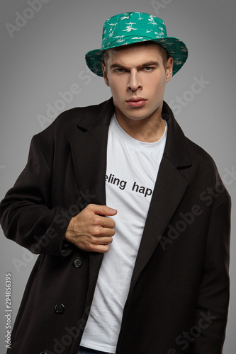 Medium close-up shot of a short-haired European man dressed in a white top with black lettering, a black jacket and a green bucket hat with white palm print.