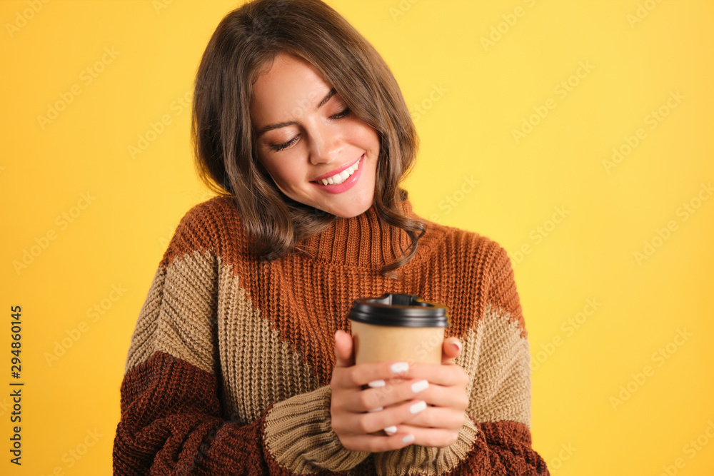 Beautiful smiling girl in sweater happily holding coffee to go over yellow background Beautiful smiling girl in sweater happily holding coffee to go over yellow background