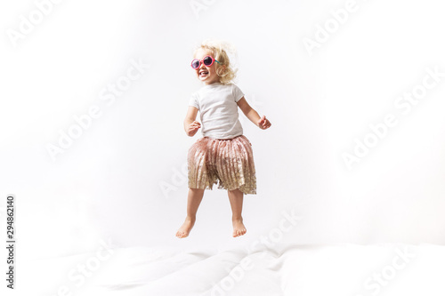 A little girl in pink sunglasses and a pink skirt jumps on a white mattress. © Aboltin