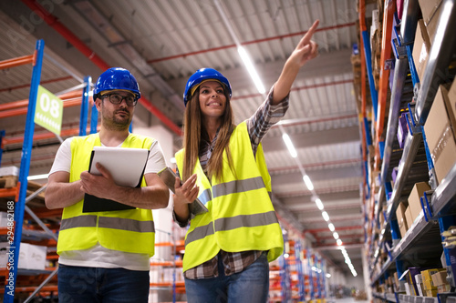 Shot of positive cheerful warehouse workers checking inventory on shelves together and controlling distribution of products in large storage area.