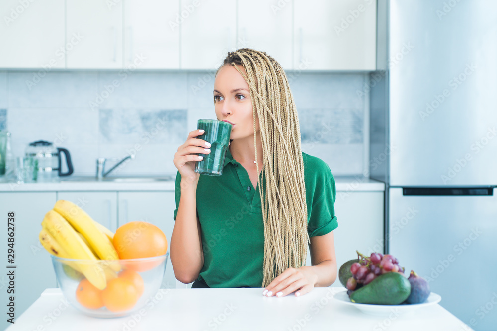 Young woman drinking green smoothie in the kitchen with fruits and vegetables. Healthy eating lifestyle. Woman drinking matcha from glass at home. Healthy life and food concept.